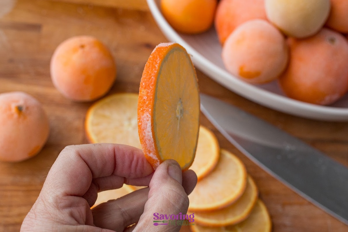 A hand holds a thin slice of orange fruit above a cutting board with more slices, a knife, and a plate of whole fruits in the background.