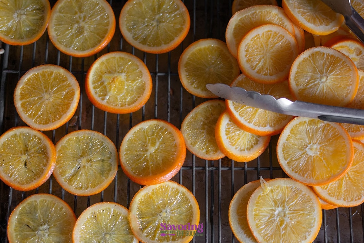 Sliced oranges drying on a wire rack, with metal tongs placed next to a pile of overlapping slices.