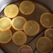 Sliced oranges and a vanilla bean simmer in a pot of liquid with visible specks of seasoning.