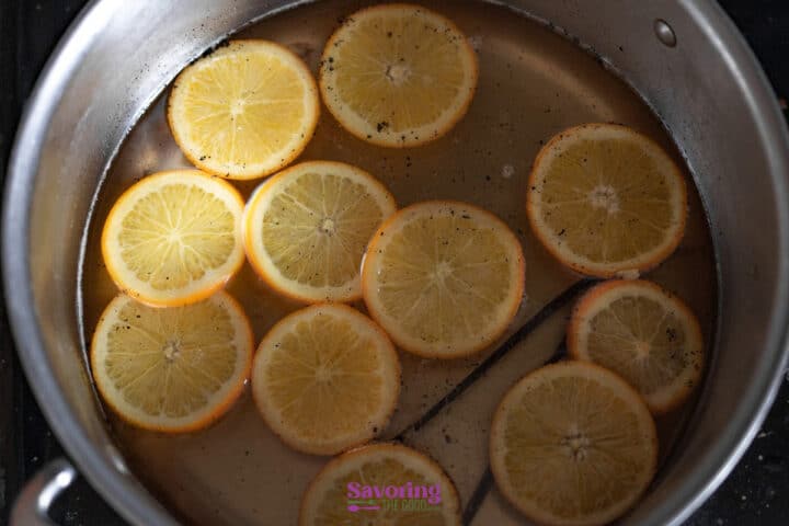Sliced oranges and a vanilla bean simmer in a pot of liquid with visible specks of seasoning.