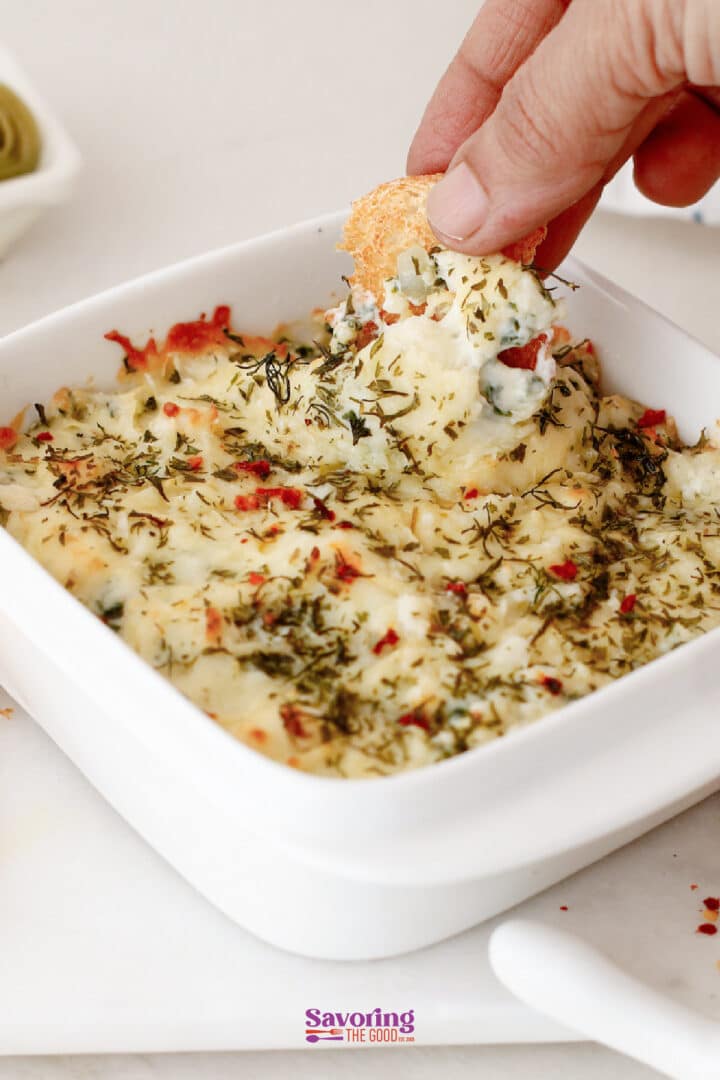 A hand dips a round cracker into a baked spinach artichoke dip topped with herbs in a white square dish.