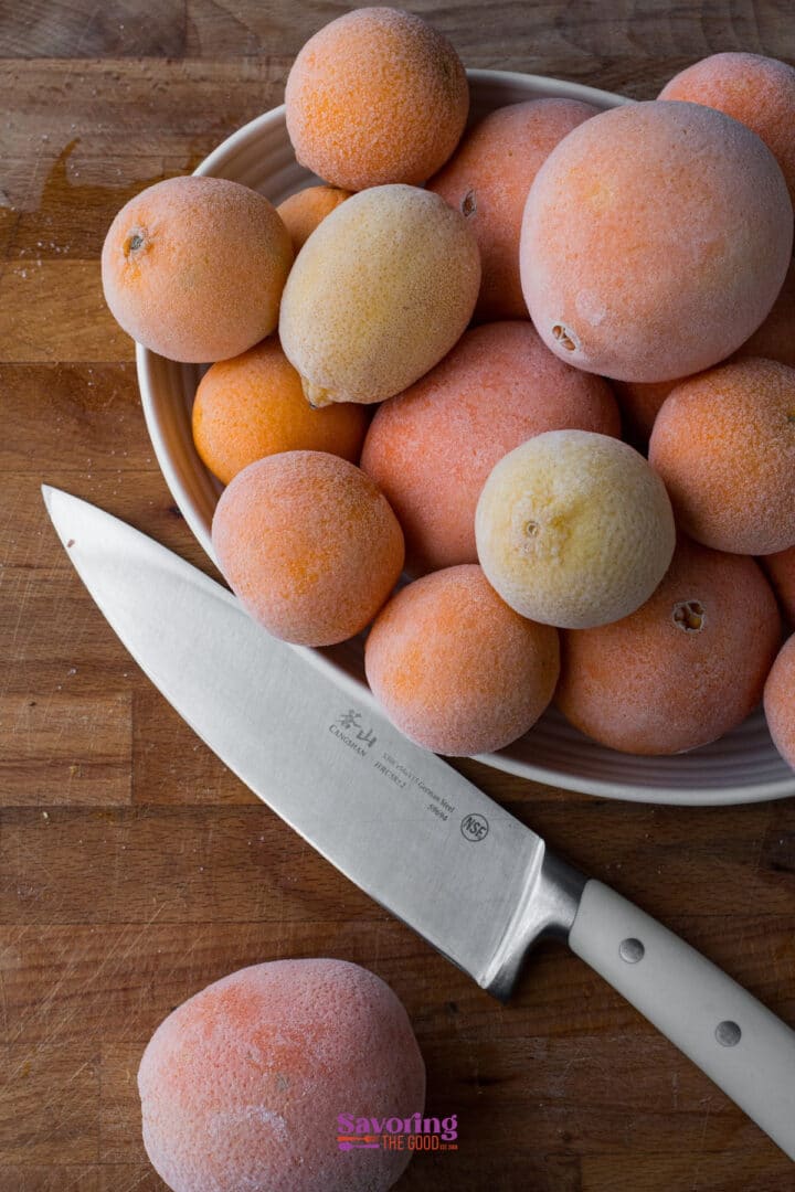 A white bowl filled with frozen lemons and oranges sits on a wooden surface next to a chef's knife.