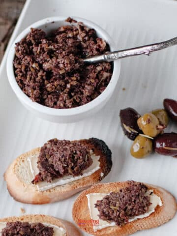 A white dish with olive tapenade, a small bunch of green and black olives, and slices of bread topped with tapenade on a rectangular plate.