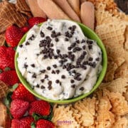 A green bowl of creamy cannoli dip topped with mini chocolate chips, surrounded by strawberries, waffle cookies, and ladyfinger biscuits on a platter.