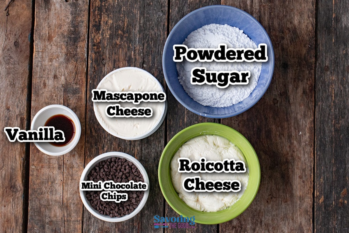 Bowls on a wooden table containing ingredients for cannoli dip-powdered sugar, mascarpone cheese, ricotta cheese, mini chocolate chips, and vanilla extract-each labeled and ready to use.