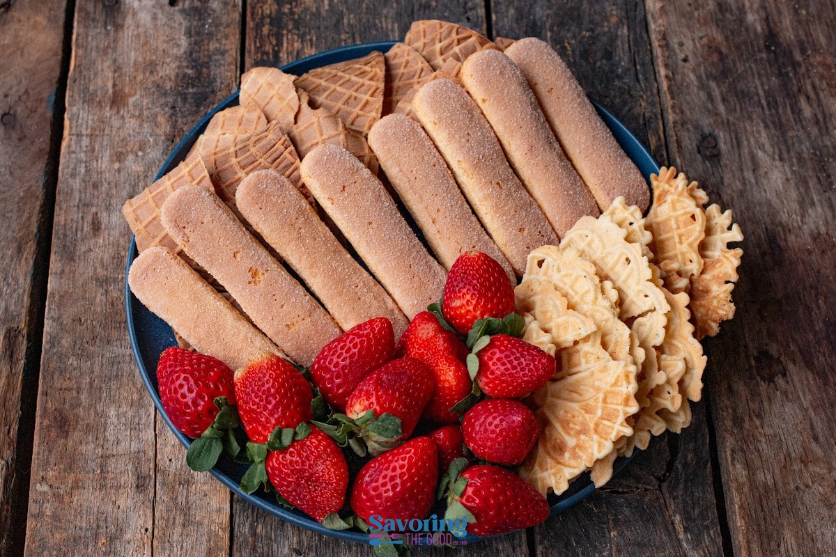 A plate with ladyfinger biscuits, waffle cookies, fresh strawberries, and creamy cannoli dip on a wooden surface.