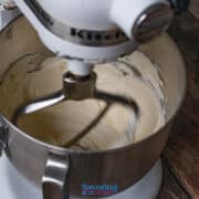 A stand mixer with a paddle attachment is blending creamy cannoli dip in a metal bowl on a wooden surface.