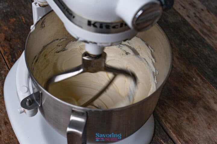 A stand mixer with a paddle attachment is blending creamy cannoli dip in a metal bowl on a wooden surface.