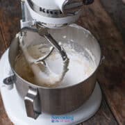 A stand mixer with a metal bowl contains partially mixed cannoli dip batter, sitting on a rustic wooden surface.