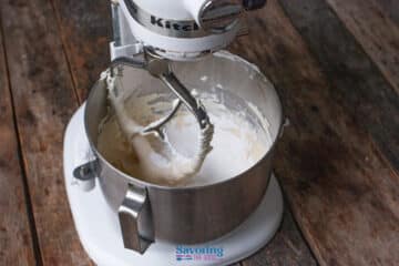 A stand mixer with a metal bowl contains partially mixed cannoli dip batter, sitting on a rustic wooden surface.