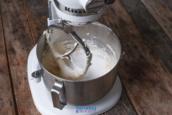 A stand mixer with a metal bowl contains partially mixed cannoli dip batter, sitting on a rustic wooden surface.