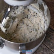 Stand mixer bowl filled with Cannoli dip mixed with chocolate chips with a paddle attachment coated in batter, on a wooden surface.