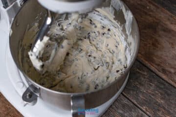 Stand mixer bowl filled with Cannoli dip mixed with chocolate chips with a paddle attachment coated in batter, on a wooden surface.