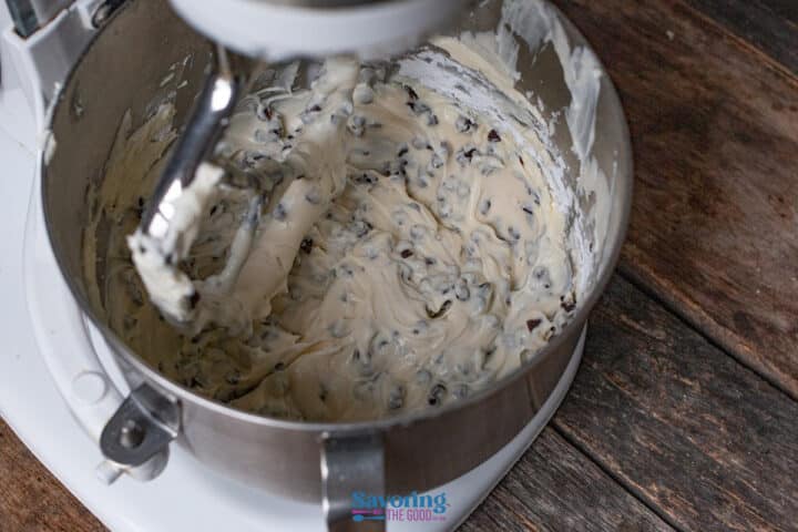 Stand mixer bowl filled with Cannoli dip mixed with chocolate chips with a paddle attachment coated in batter, on a wooden surface.
