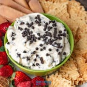 A green bowl filled with creamy cannoli dip topped with mini chocolate chips, surrounded by strawberries, waffle cookies, and ladyfingers on a platter.