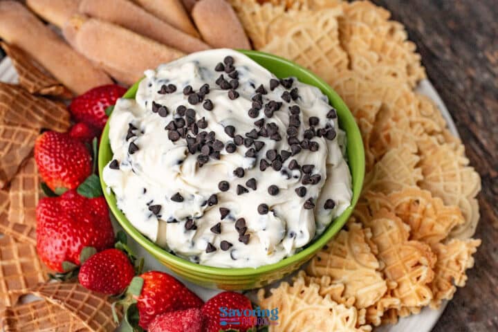 A green bowl filled with creamy cannoli dip topped with mini chocolate chips, surrounded by strawberries, waffle cookies, and ladyfingers on a platter.