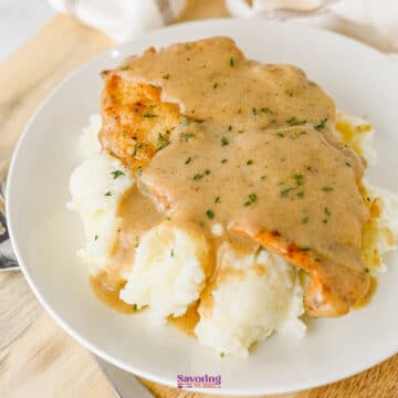 A plate of mashed potatoes topped with a breaded, smothered chicken cutlet and covered in brown gravy, garnished with chopped herbs.