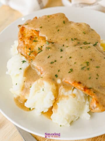 A plate of mashed potatoes topped with a breaded, smothered chicken cutlet and covered in brown gravy, garnished with chopped herbs.