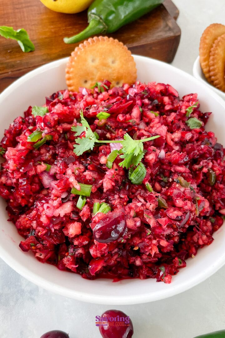 A white bowl filled with cranberry salsa topped with cilantro, with a round cracker on the rim and more crackers and ingredients in the background.