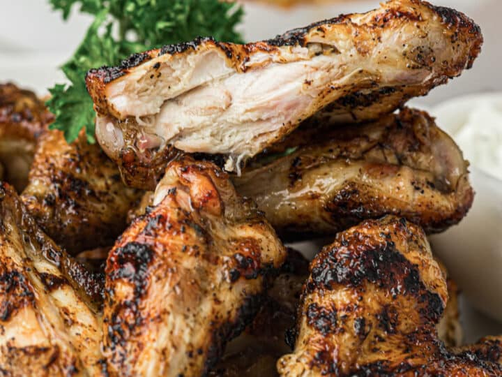 A close-up of grilled chicken wings stacked on a plate, garnished with parsley, with a side of dipping sauce in the background.