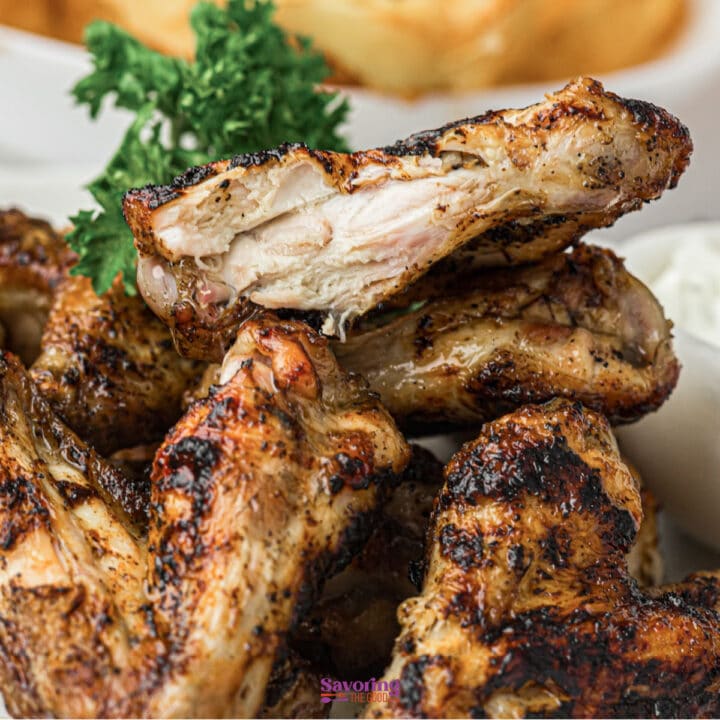 A close-up of grilled chicken wings stacked on a plate, garnished with parsley, with a side of dipping sauce in the background.