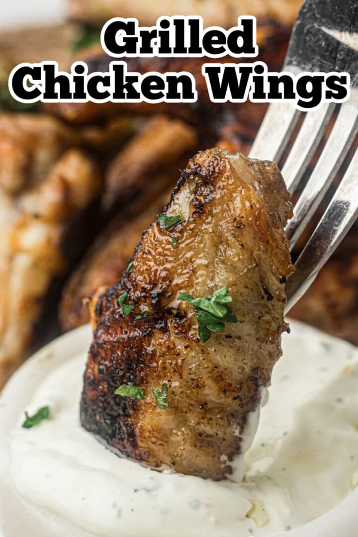 A grilled chicken wing held by a fork is dipped in a bowl of white sauce, with more grilled wings visible in the background.
