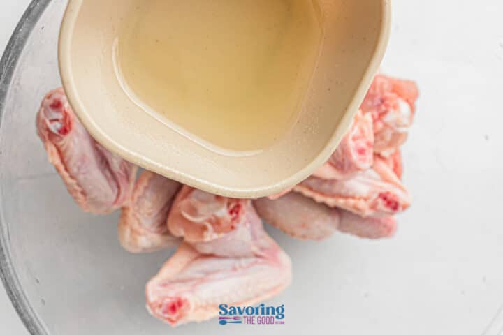 A bowl of raw chicken wings in a glass bowl, with a separate bowl of clear liquid being poured over them.