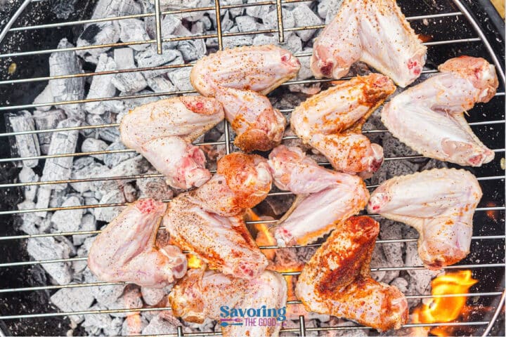Raw, seasoned chicken wings coated in a flavorful dry rub are arranged on a metal grill over hot charcoal and flames, ready to become delicious grilled chicken wings.