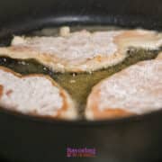 Three pieces of lightly floured chicken are being pan-fried in oil in a black skillet.