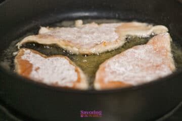 Three pieces of lightly floured chicken are being pan-fried in oil in a black skillet.