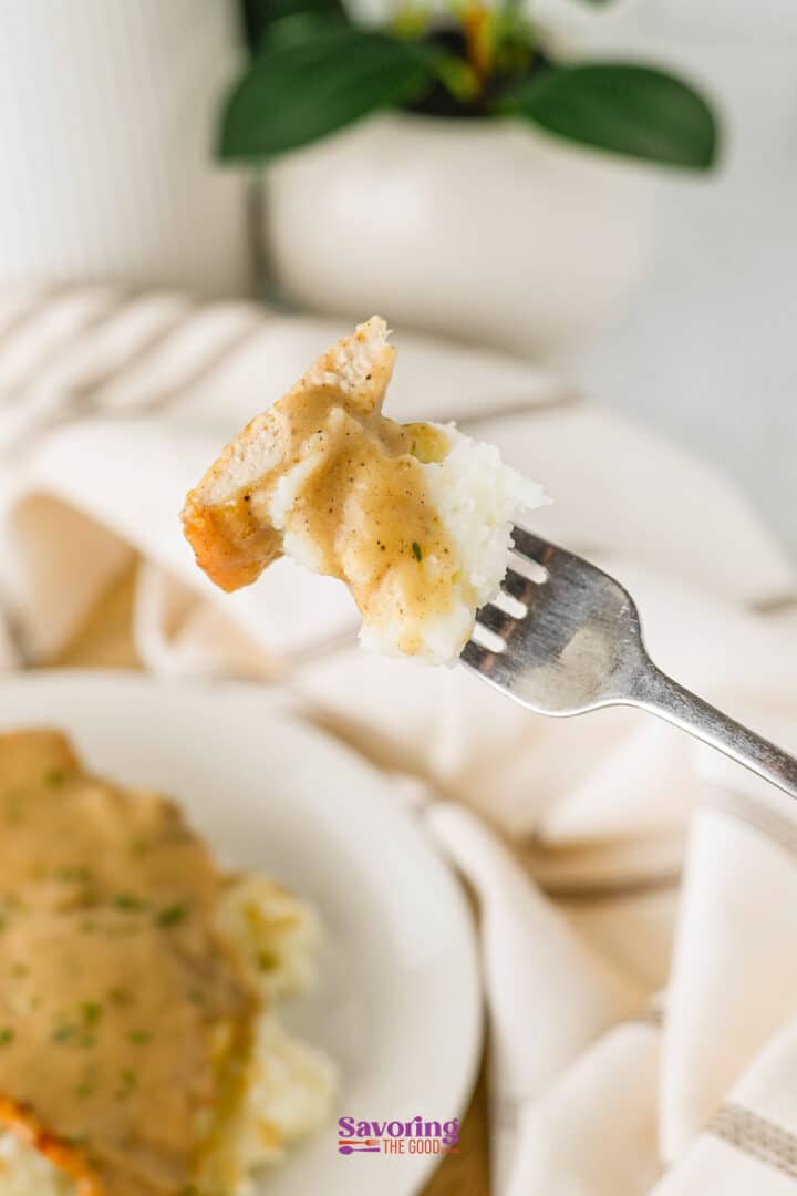 A fork holds a bite of smothered chicken and mashed potatoes with gravy, with a plate of food and a potted plant blurred in the background.