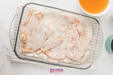 Raw chicken cutlets coated in flour are arranged in a glass baking dish on a striped towel, with orange liquid and a shaker nearby.