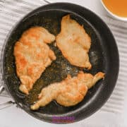 Three pieces of breaded chicken are being pan-fried in a black skillet with oil, placed on a striped cloth next to a bowl of liquid on a white surface.
