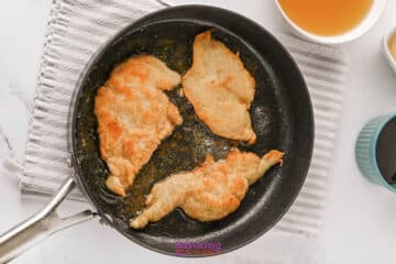 Three pieces of breaded chicken are being pan-fried in a black skillet with oil, placed on a striped cloth next to a bowl of liquid on a white surface.
