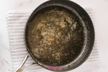 A black saucepan with browned butter residue sits on a white textured cloth on a marble surface.