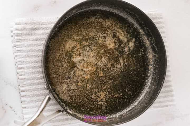 A black saucepan with browned butter residue sits on a white textured cloth on a marble surface.