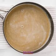 A saucepan containing a light brown liquid sits on a white striped towel atop a marble surface.