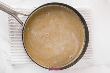 A saucepan containing a light brown liquid sits on a white striped towel atop a marble surface.
