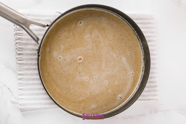 A saucepan containing a light brown liquid sits on a white striped towel atop a marble surface.