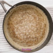 A black pan containing browned butter on a white textured towel, roux for smothered chicken, viewed from above on a white surface.