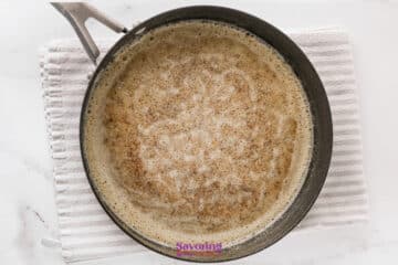 A black pan containing browned butter on a white textured towel, roux for smothered chicken, viewed from above on a white surface.