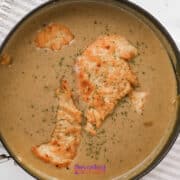 A saucepan with browned chicken cutlets in a creamy brown gravy, garnished with herbs, on a striped towel.