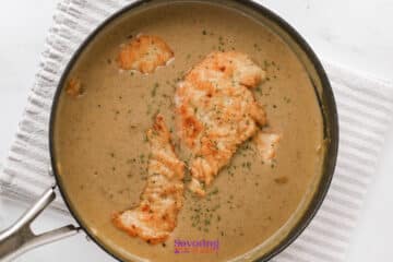 A saucepan with browned chicken cutlets in a creamy brown gravy, garnished with herbs, on a striped towel.