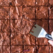 A metal spatula lifts a single chocolate brownie from a tray of evenly cut sheet pan brownies.