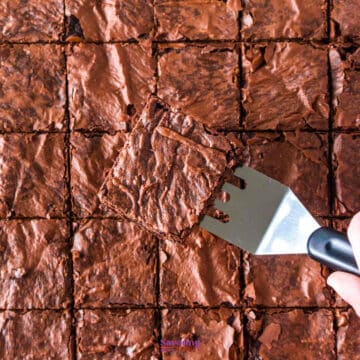 A metal spatula lifts a single chocolate brownie from a tray of evenly cut sheet pan brownies.