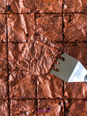 A metal spatula lifts a single chocolate brownie from a tray of evenly cut sheet pan brownies.
