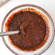 A glass bowl filled with chocolate cake batter being stirred with a metal spoon on a white surface.