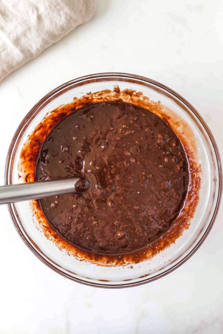 A glass bowl filled with chocolate cake batter being stirred with a metal spoon on a white surface.