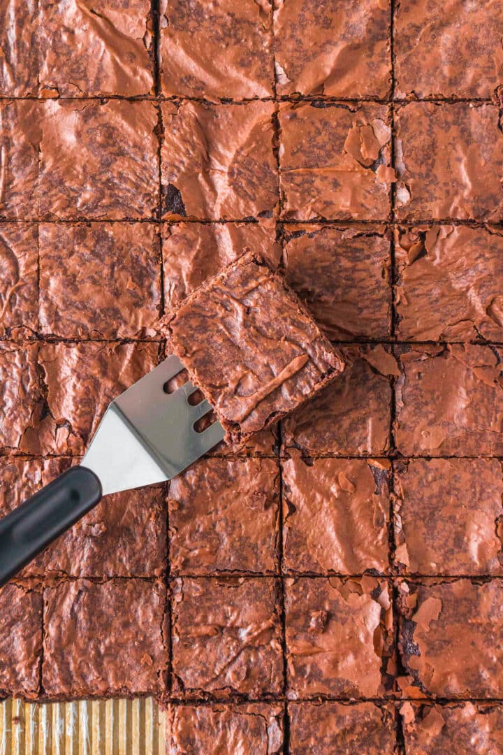 A spatula lifts one square brownie from a batch of evenly cut Sheet Pan Brownies with a shiny, cracked top.