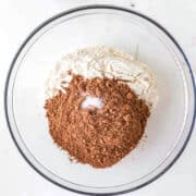 A glass bowl with flour, cocoa powder, and salt on a white surface, with small bowls of chocolate chips and chocolate shavings nearby.